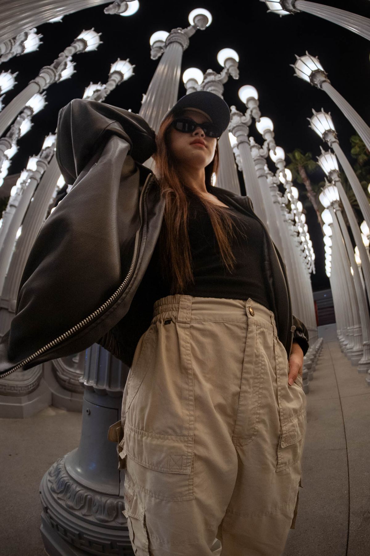 Woman in a cap and oversized jacket beneath a canopy of street lamps
