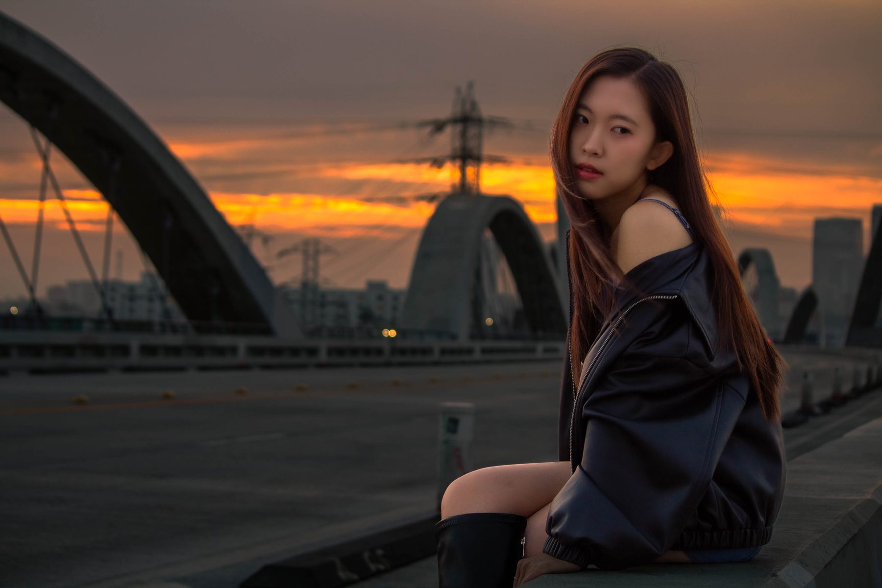 Woman in a leather jacket seated on a bridge at sunset